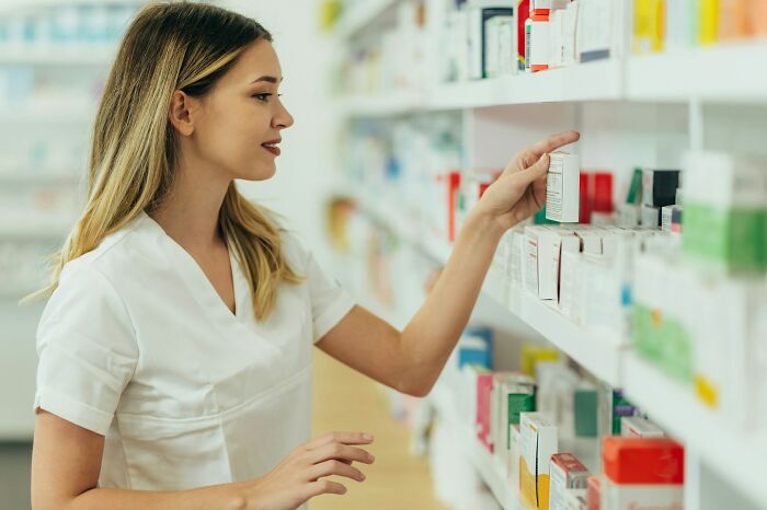 Pharmacist in white uniform organizing medicine boxes on shelves illustrating jobs that shouldn’t be earning as much as they do.