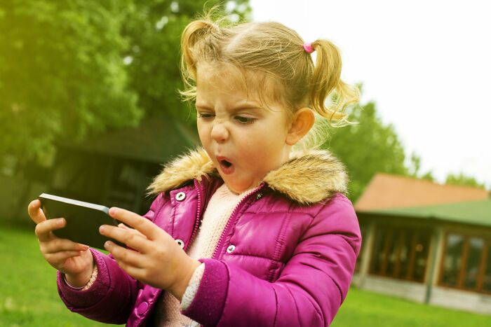 Young girl in a purple jacket outside, showing a surprised reaction while looking at a smartphone, capturing boomer opinions.