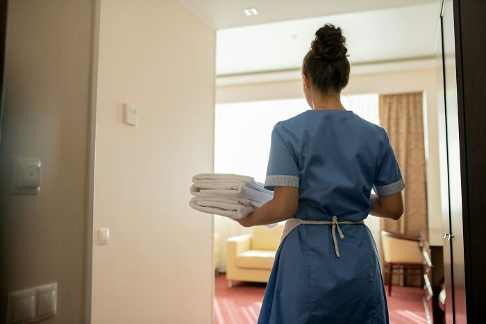 Housekeeper in blue uniform carrying folded towels entering a room revealing a company secret workplace scene