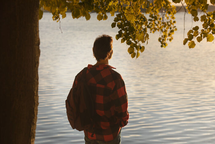 A man in a red plaid shirt with a backpack stands by the water, reflecting on life after relationship mistakes.