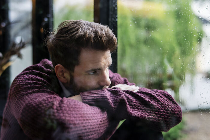 Man in a purple sweater looking out a rainy window, reflecting on life after losing an amazing partner.