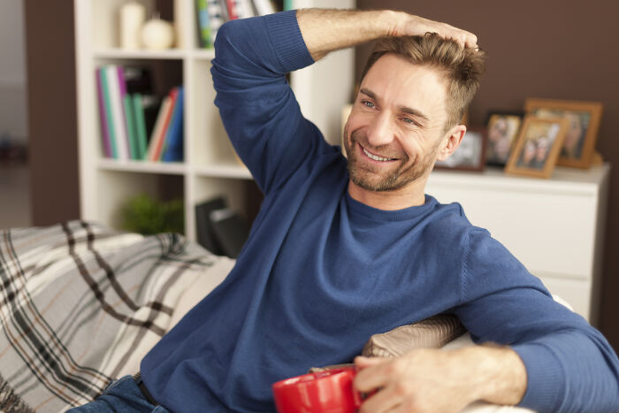 Smiling man in a blue sweater sitting on a couch holding a red mug, reflecting on life changes after relationship mistakes.