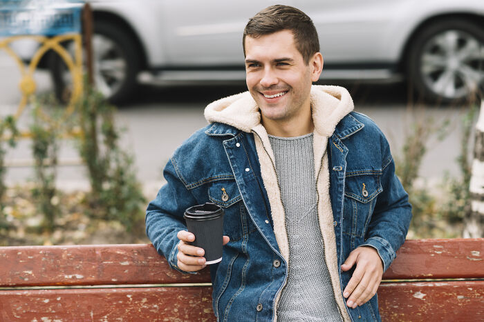 Smiling man sitting on a bench outdoors, holding a coffee cup, reflecting on life changes after relationship challenges.