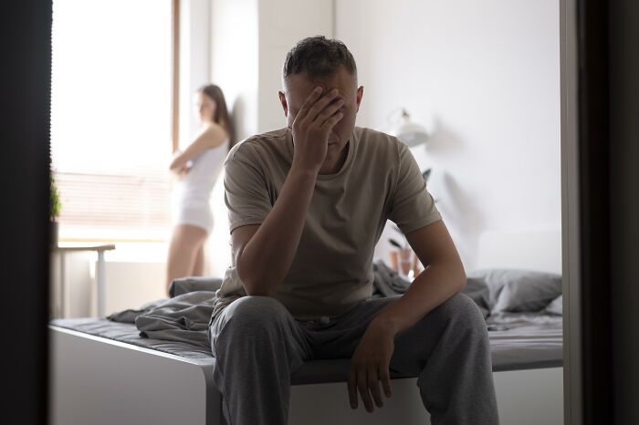 Man holding his face in distress on bed while woman stands upset in background, reflecting life after ruining relationship.