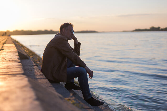 A man sitting alone by the water at sunset, reflecting on life after he ruined things with his partner.