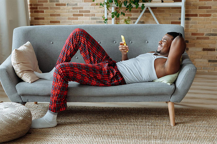 Man in red plaid pants relaxing on a gray sofa, smiling while looking at his phone in a cozy living room setting.