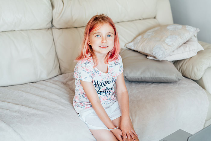 Young girl with pink-dyed hair smiling while sitting on a beige couch after babysitter dyes girls&rsquo; hair pink.