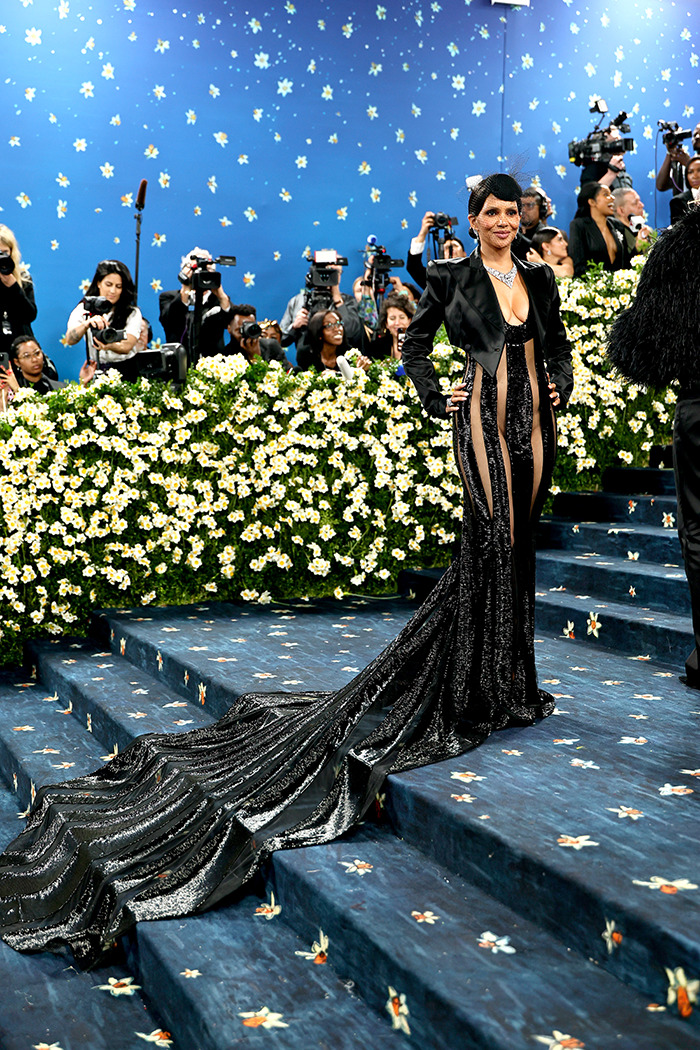 Halle Berry posing on the Met Gala stairs in a sheer, underwear-free black dress with a long train and floral backdrop.