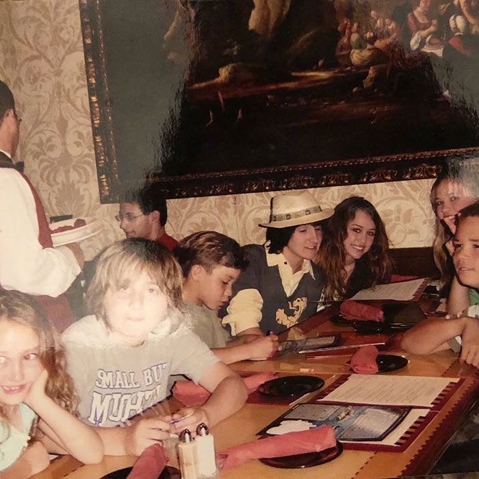 Noah Cyrus in a casual setting with friends, sporting a relaxed look amid a group at a dining table.