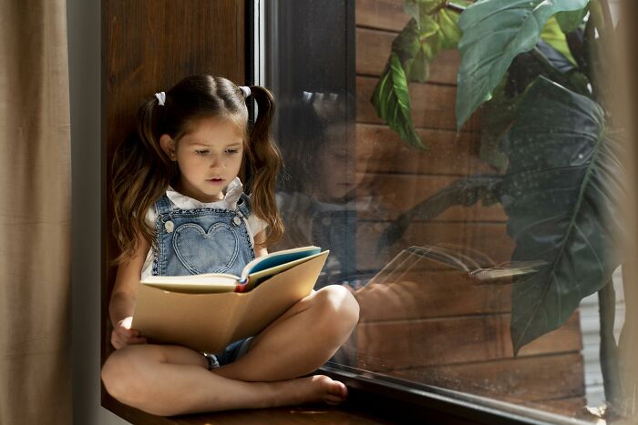 Young girl reading a book by the window, illustrating childhood habits and possible red flags in behavior.