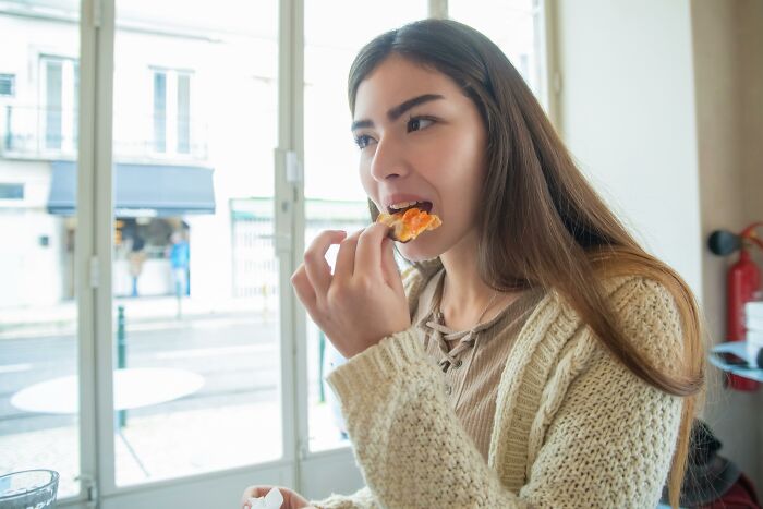 Young woman eating a snack indoors near a window, illustrating childhood habits and potential red flags behavior.
