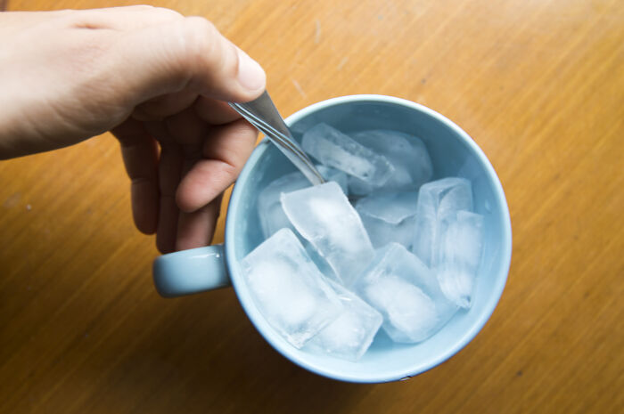 Hand holding a spoon stirring ice cubes in a blue cup on a wooden surface, highlighting attention-seeking behaviors.