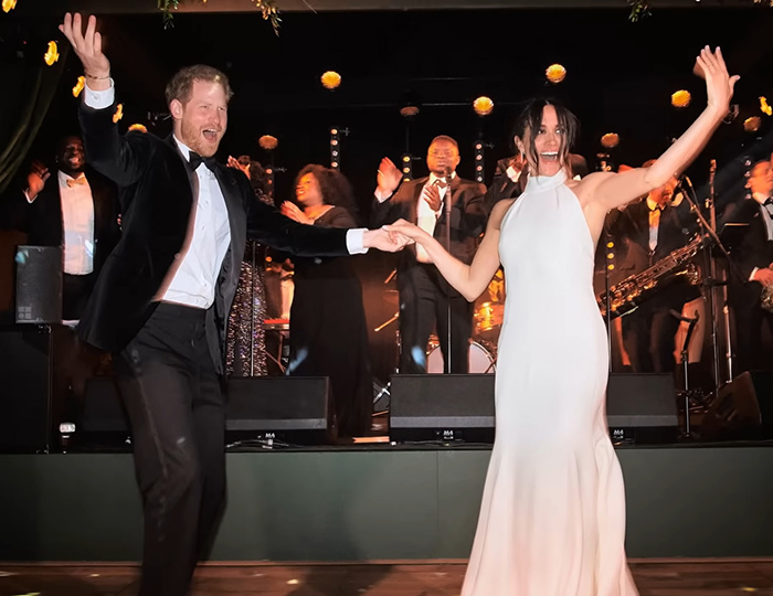 Prince Harry and Meghan Markle dancing joyfully at an event with a live band performing on stage behind them.