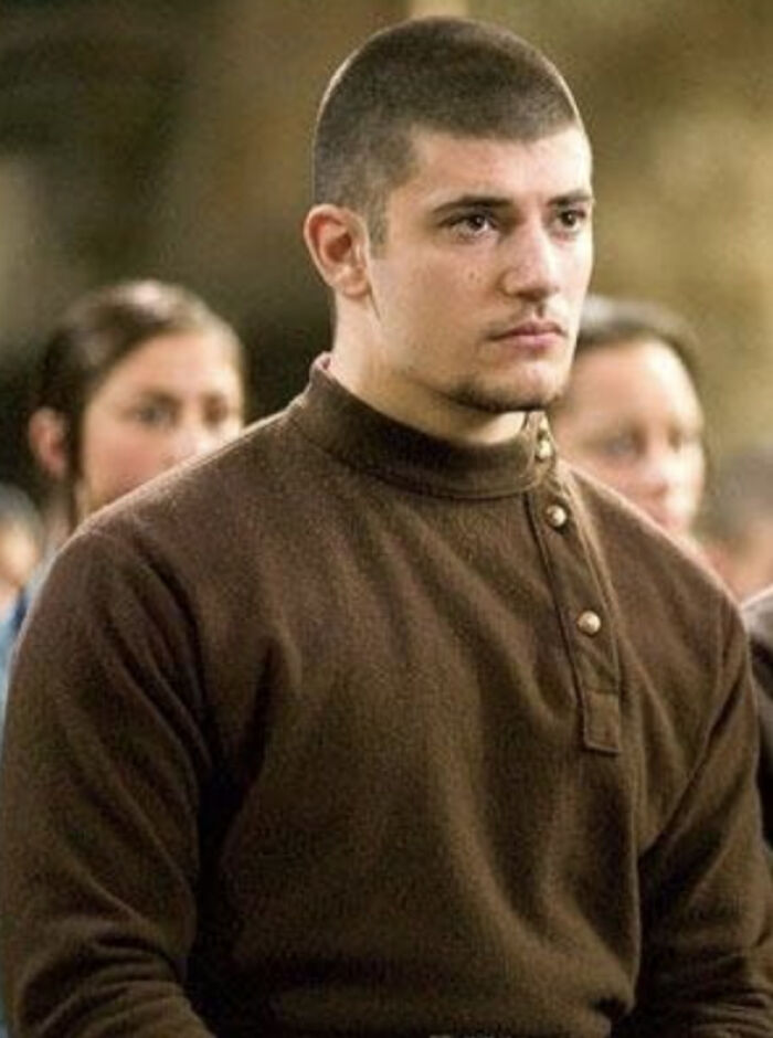 Actor who played Viktor Krum in Harry Potter looking serious in a brown sweater during a film scene with blurred people in background