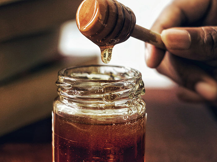 Close-up of honey dripping from a wooden dipper into a jar, illustrating ‘healthy’ foods you might want to skip eating.