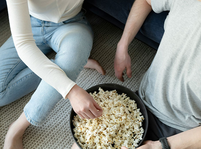 Two people sitting on the floor sharing a large bowl of popcorn, illustrating healthy foods to skip eating advice.
