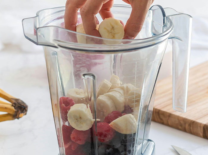Hand placing banana slices into blender with raspberries and blueberries to prepare healthy food ingredients.