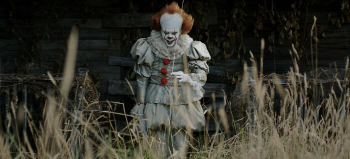 Eerie clown standing in tall grass near an old wooden shack, illustrating unconventional crushes people actually have.