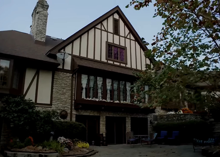 Suspected serial killer's farm with Tudor-style house surrounded by trees and outdoor seating in dim daylight.