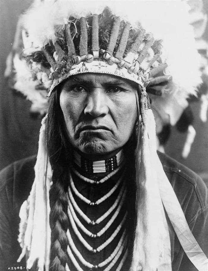 Portrait of a Native American man wearing traditional headdress and bead necklace in a powerful historic portrait.