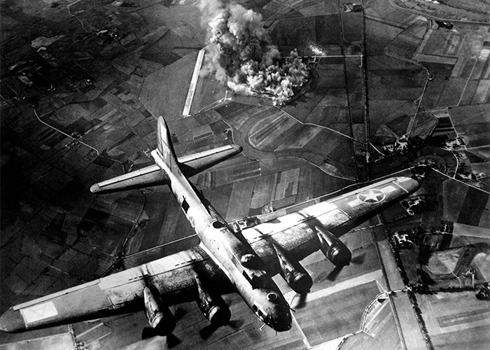 Black and white aerial photo of a bomber plane over farmland with smoke from an explosion in historical events.
