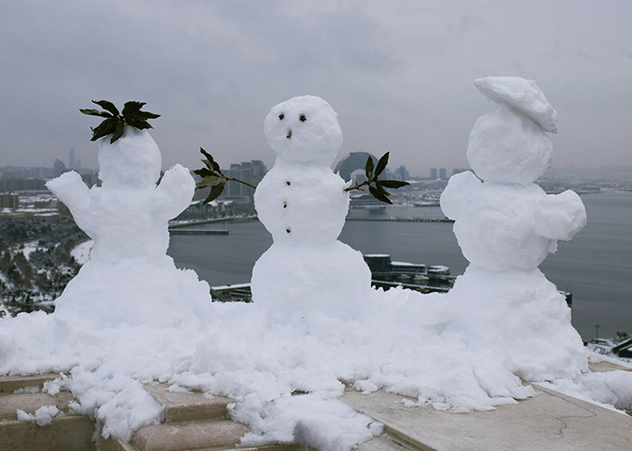 Three snowmen with leafy arms standing on a snowy ledge overlooking a cityscape on a winter day historical events