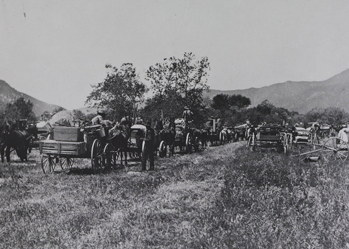 Black and white photo of a historical event showing soldiers and horse-drawn wagons in a rural setting.