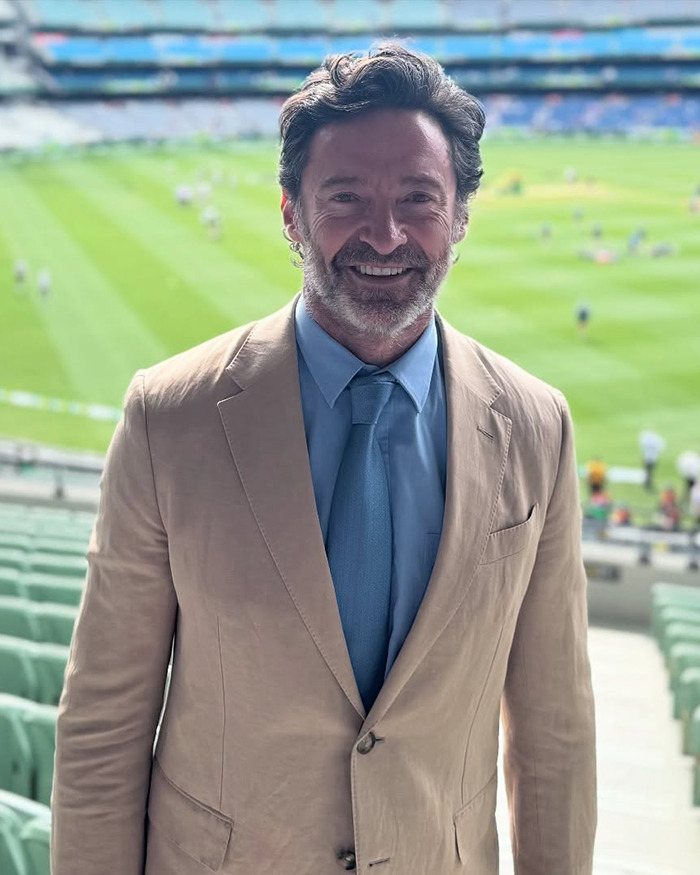 Hugh Jackman smiling in a beige suit and blue tie at a sports stadium, related to his romance rough patch news. Hugh Jackman smiling in a beige suit and blue tie at a sports stadium, related to his romance rough patch news.