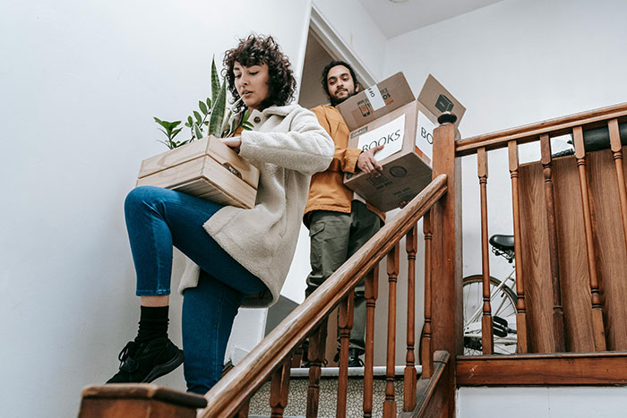 Woman carrying a plant and man holding moving boxes while descending stairs during cross-country move.