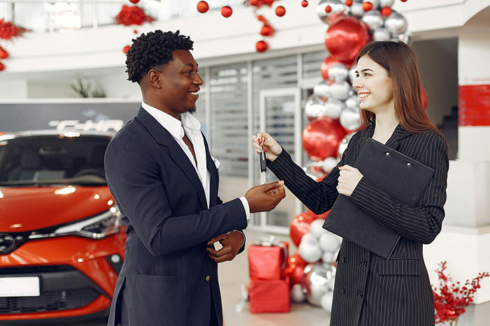 Woman sells everything for cross-country move, handing car keys to a man in a festive dealership setting.