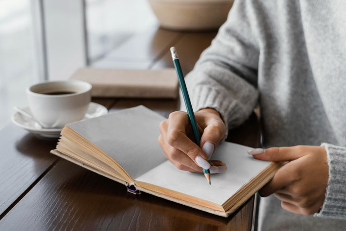 Woman writing in a notebook at a table with a coffee cup, illustrating dynamics of undermining a wife&rsquo;s job in front of others.
