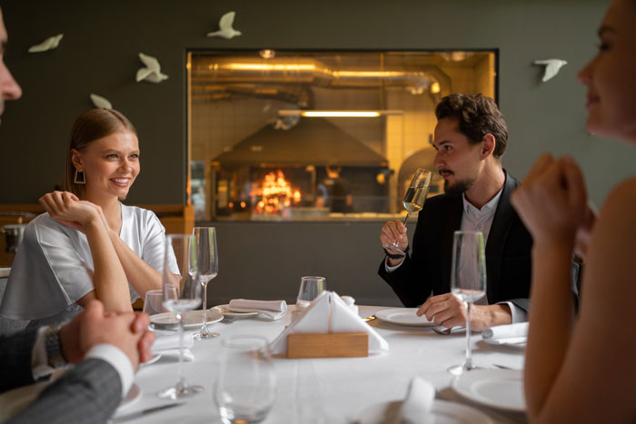 Couple dining with others at a restaurant, man appearing defensive while woman smiles during conversation about work.