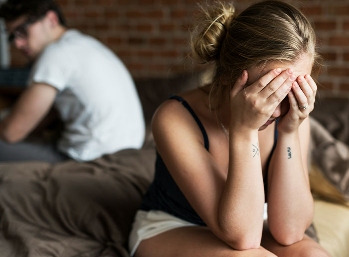 Woman sits on bed covering face in shock, husband turns away, capturing reaction to pregnancy test surprise. Woman sits on bed covering face in shock, husband turns away, capturing reaction to pregnancy test surprise.