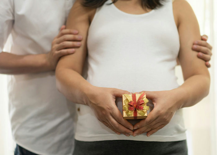 Pregnant woman holding a small gift box with her husband standing behind, symbolizing pregnancy test surprise gift. Pregnant woman holding a small gift box with her husband standing behind, symbolizing pregnancy test surprise gift.