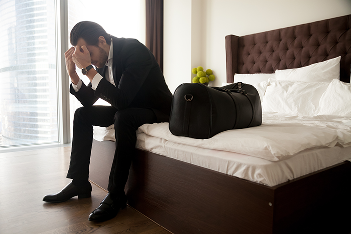 Man in suit sitting on bed with travel bag, looking distressed after wife's cancer diagnosis impacts plans for biological kids.