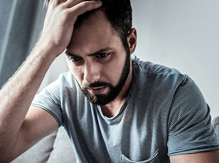 Man with beard in striped shirt looking worried and stressed while sitting indoors, contemplating an irresponsible manly purchase.