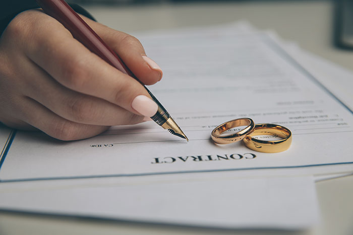 Close-up of a hand signing a divorce contract with wedding rings nearby representing post-partum custody conflict. Close-up of a hand signing a divorce contract with wedding rings nearby representing post-partum custody conflict.