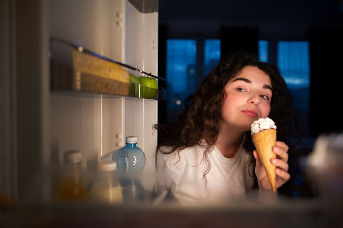 Woman eating ice cream at night near open fridge, illustrating parenting conflict over nightly ice cream habits. Woman eating ice cream at night near open fridge, illustrating parenting conflict over nightly ice cream habits.