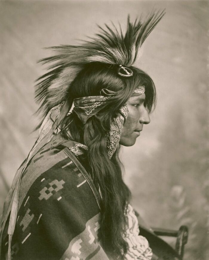 Native American man in traditional attire with feathered headdress and patterned blanket in a powerful historical portrait.
