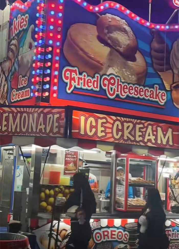 County fair food stand with bright lights advertising fried cheesecake, lemonade, ice cream, and fried Oreos at night.