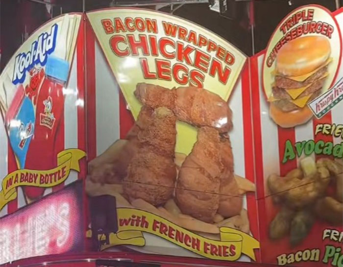 Fair food stand featuring bacon wrapped chicken legs with french fries and a triple cheeseburger at a county fair.