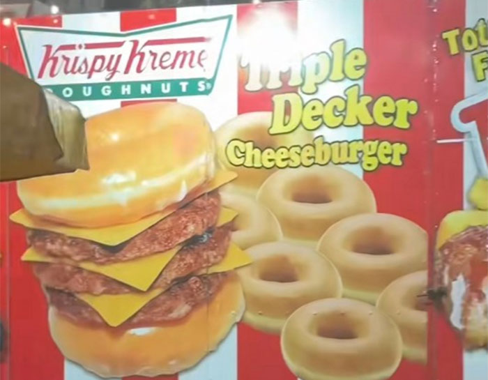Triple decker cheeseburger with Krispy Kreme doughnuts at a county fair food stall display.