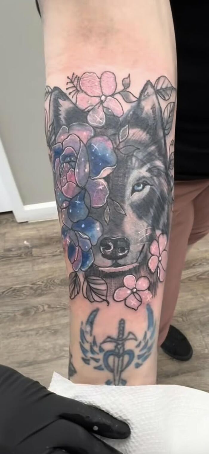Tattoo of a wolf partially covered with colorful flowers on a forearm, showcasing an attempt to look cool but with permanent mistakes.