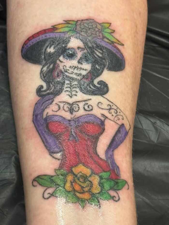 Tattoo of a colorful skeleton woman in a red dress and hat, showcasing a cool inked design with a permanent mistake visible.