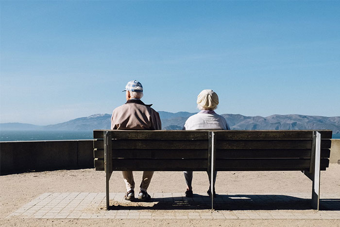 Two elderly people sitting on a bench overlooking mountains and the ocean, reflecting on changes in humanity.