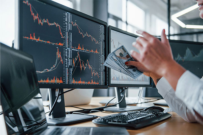 Person holding US dollar bills while monitoring financial charts on multiple computer screens illustrating future technology impact