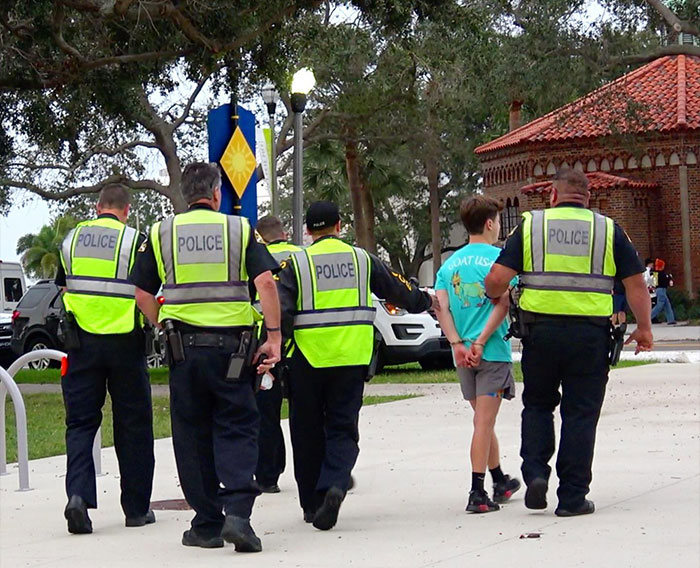 Police officers escorting Jack Doherty in handcuffs on a sidewalk near a park area with trees and a brick building.