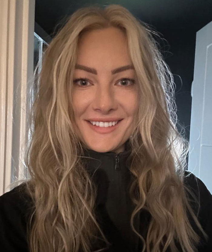 Smiling woman with long blonde hair, an experienced skydiver with over 400 flights, wearing a black jacket indoors.