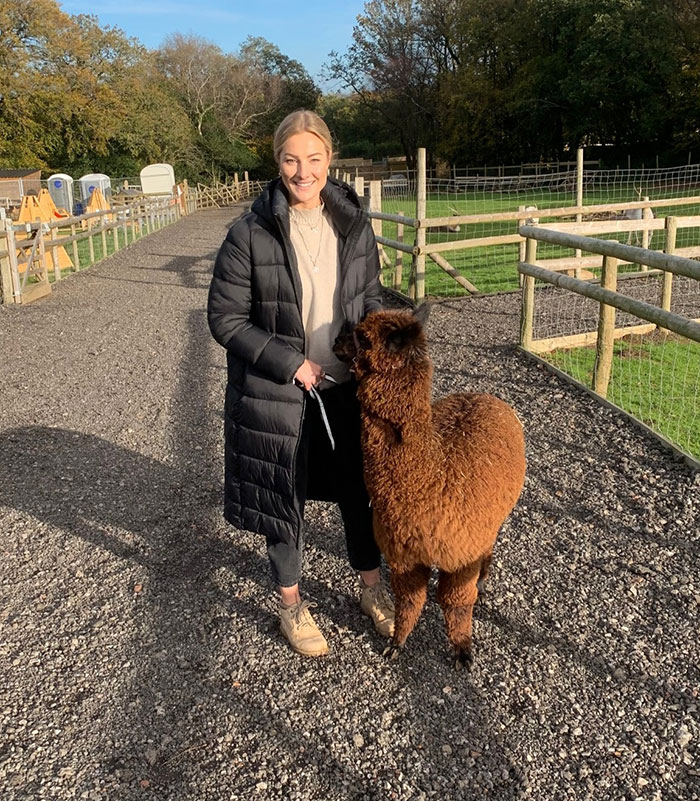 Woman with alpaca on a farm path, representing an experienced skydiver with over 400 flights in a casual outdoor setting.