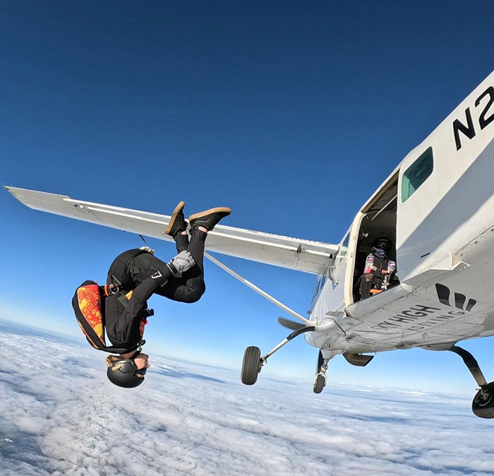 Experienced skydiver wearing helmet and gear, performing a flip while jumping from a small plane above the clouds.